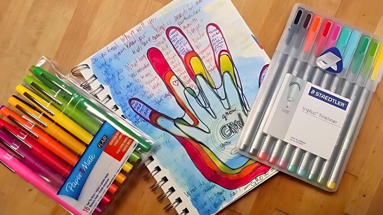 Colorful markers and pens next to an art journal with hand tracings on the page.