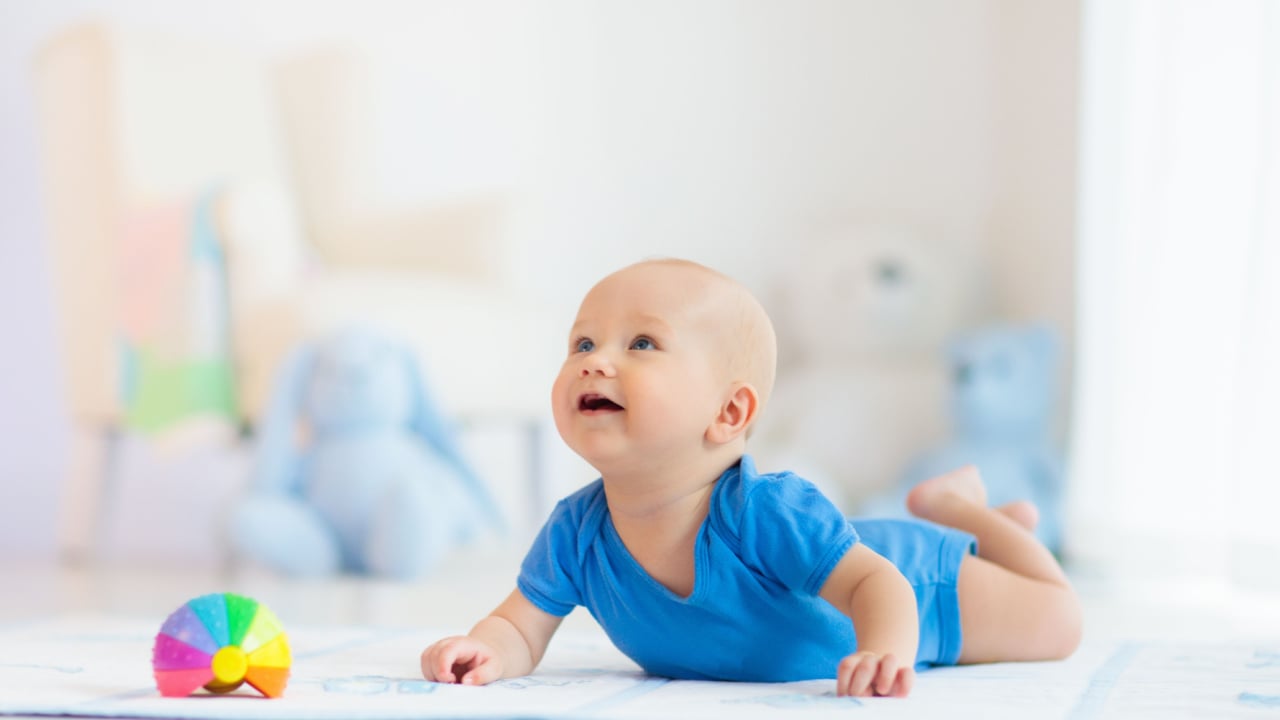 Adorable baby boy learning to crawl and playing with colorful rainbow ball toy in white sunny bedroom.