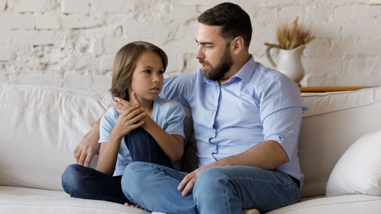 Dad sits on couch talking with son.