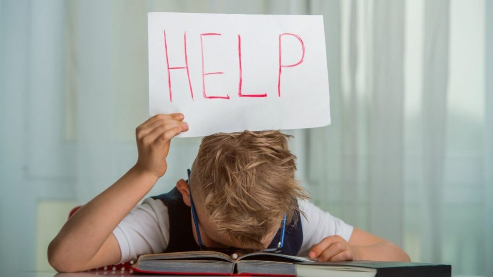 A child at a desk with head down holds a help sign over his head