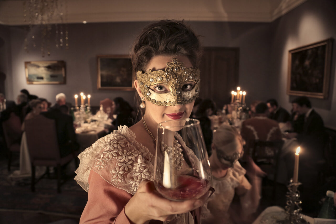 Woman in a carnival mask holds up a glass of wine.