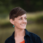 Alissa Zorn stands near a pond with an orange shirt on wearing a black button down over that.